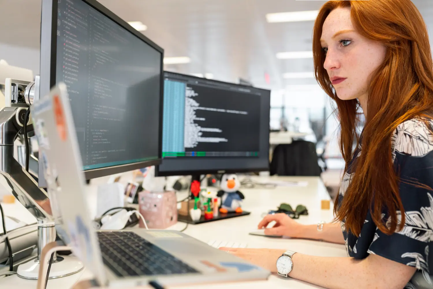 A woman working at an office desk with three screens with code.